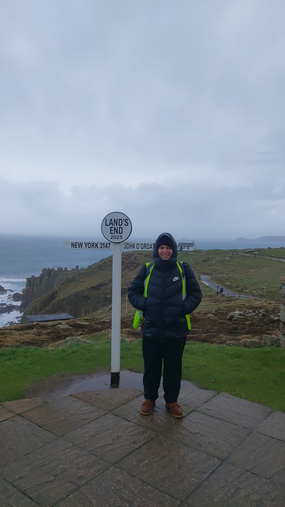 Gavin Cameron in a thick jacket, standing beside the signpost at Land's End.