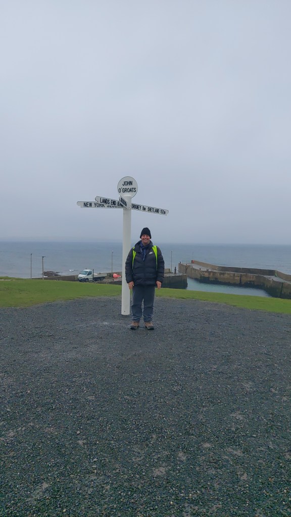 Gavin Cameron in a thick jacket, standing beside the signpost at John O'Groats.