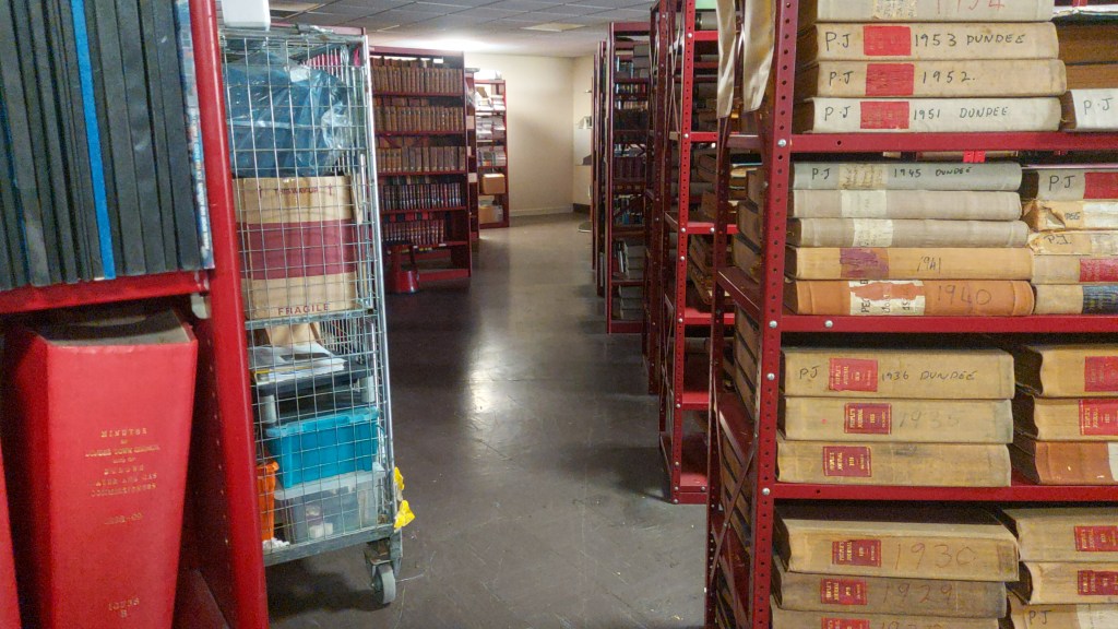 A narrow aisle lined with red shelves filled with historical documents and bound books at the Central Library, with a cart containing various bound materials in the foreground.