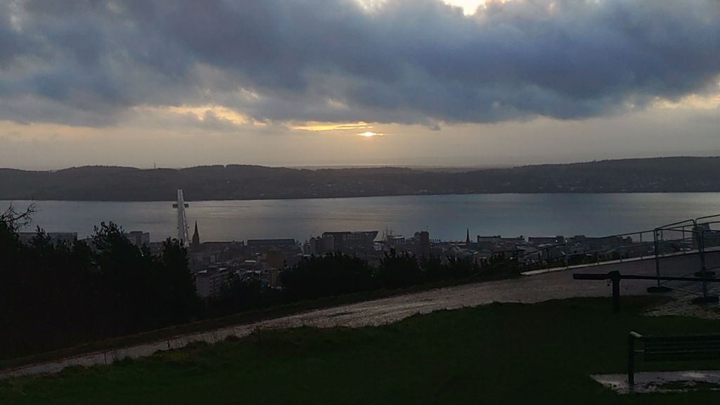 Winter solstice sunrise as seen from the Law Hill in Dundee. There is thick cloud cover partially obscuring the sun. The sky glows softly behind dramatic clouds, casting muted light across the water and buildings. A bridge spans the river, and a bench in the foreground sits beside a damp path and grassy verge.